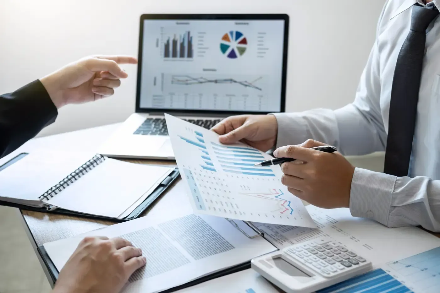 Zwei Personen sitzen an einem Schreibtisch und prüfen Finanzdaten. Eine zeigt auf einen Bildschirm mit Diagrammen, die andere hält einen Bericht in der Hand.