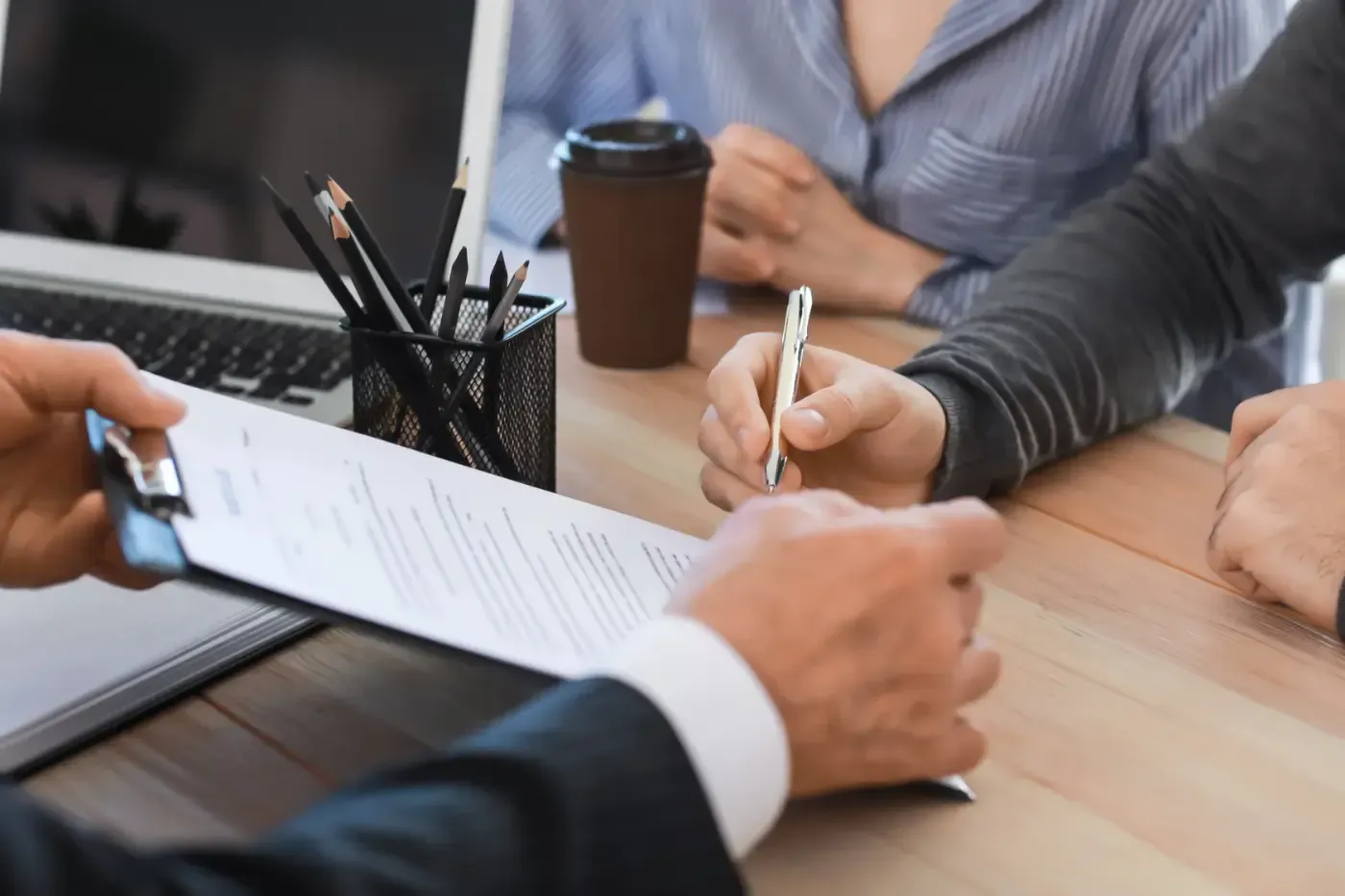 Eine Person unterschreibt ein Dokument an einem Tisch, der von jemandem im Anzug gehalten wird. Eine weitere Person sitzt daneben. Im Hintergrund sind Kaffee und ein Laptop zu sehen.
