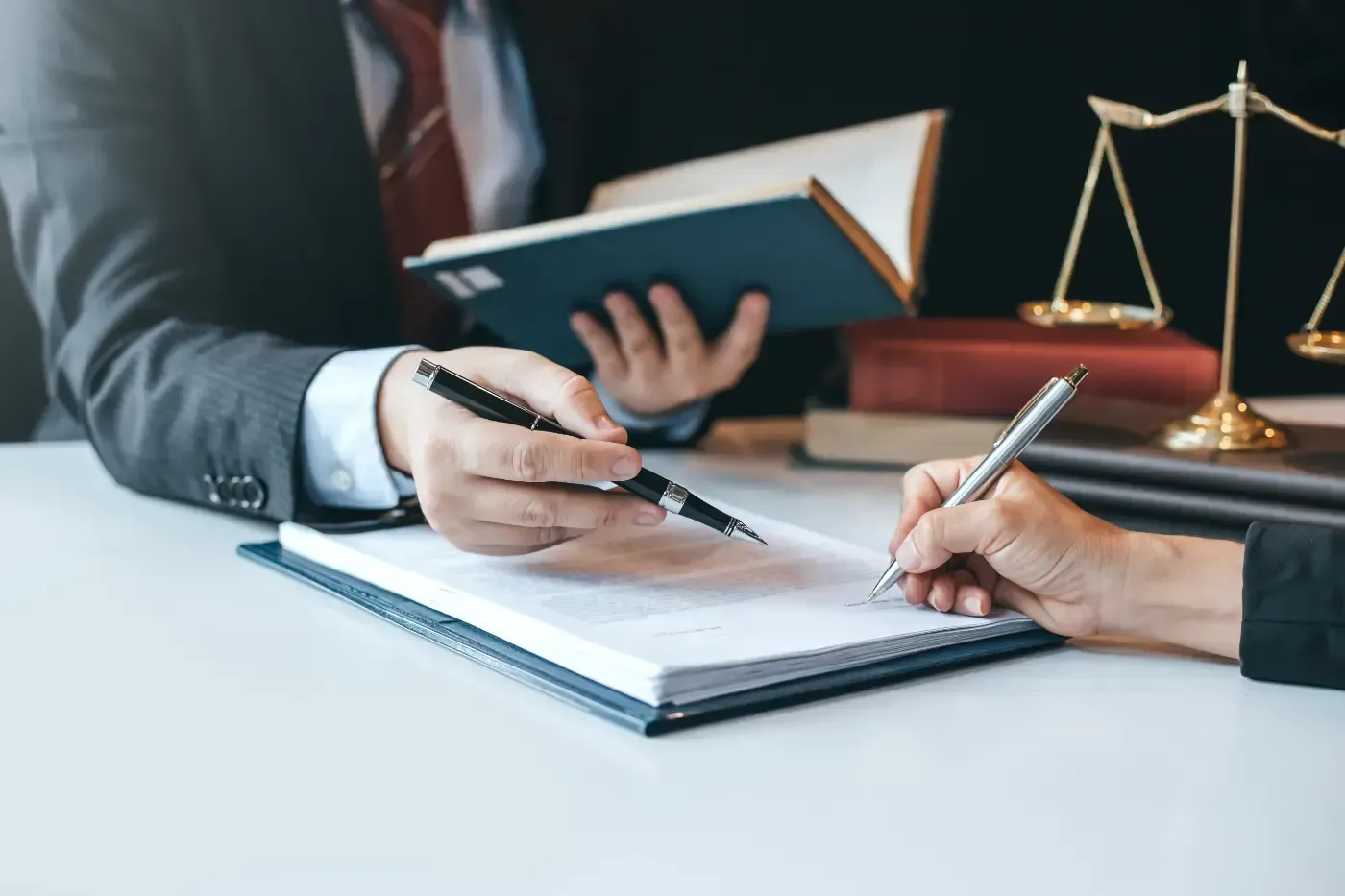 Zwei Personen an einem Schreibtisch, eine zeigt auf ein Dokument, während die andere unterschreibt. Im Hintergrund eine Justitia-Waage.