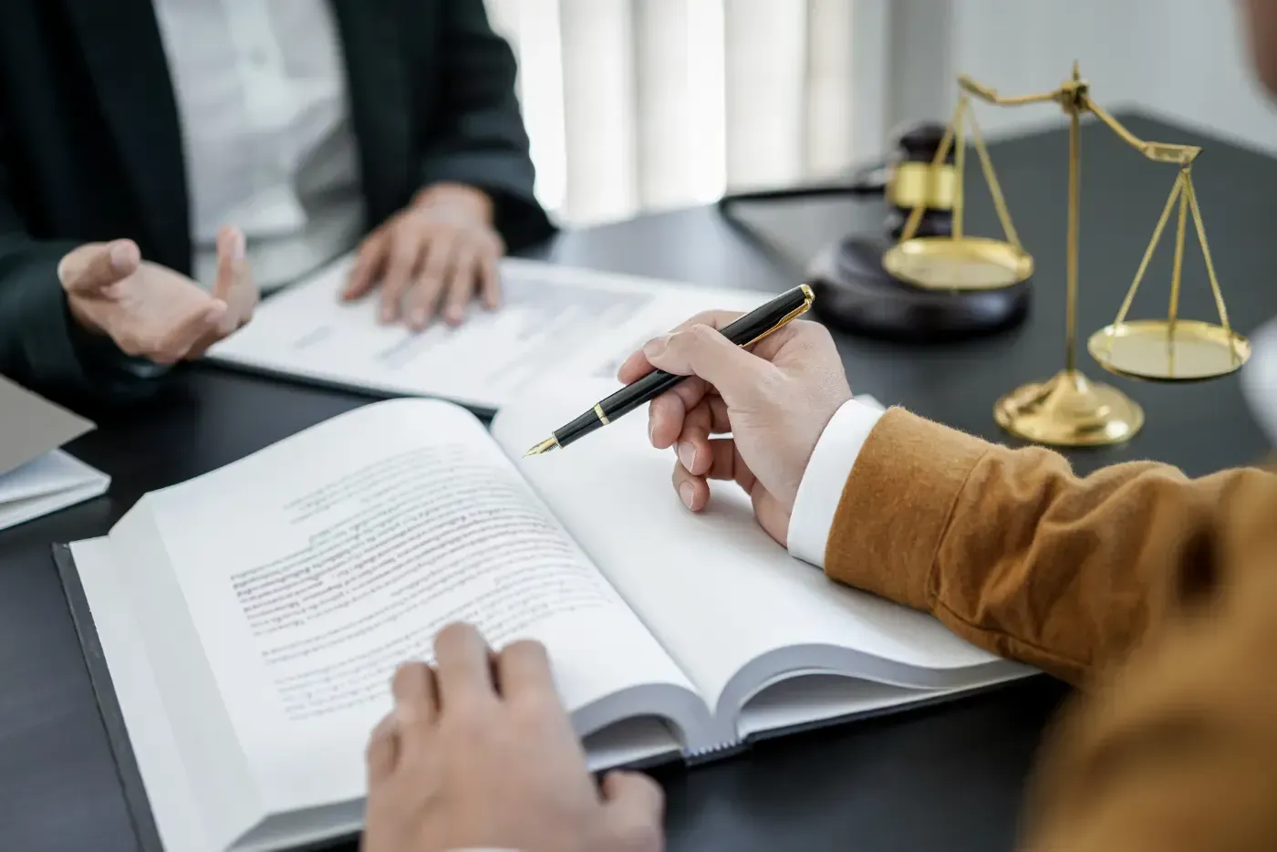 Eine Person deutet mit einem Stift auf ein aufgeschlagenes Gesetzbuch, vor einer anderen Person, die auf ein Dokument deutet. Waage und Hammer sind im Hintergrund zu sehen.