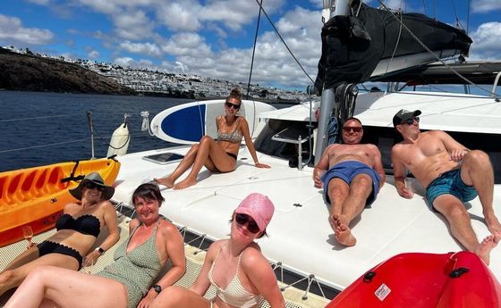 People relaxing on a sailboat deck in swimwear on a sunny day, with blue water and rocky coastline.