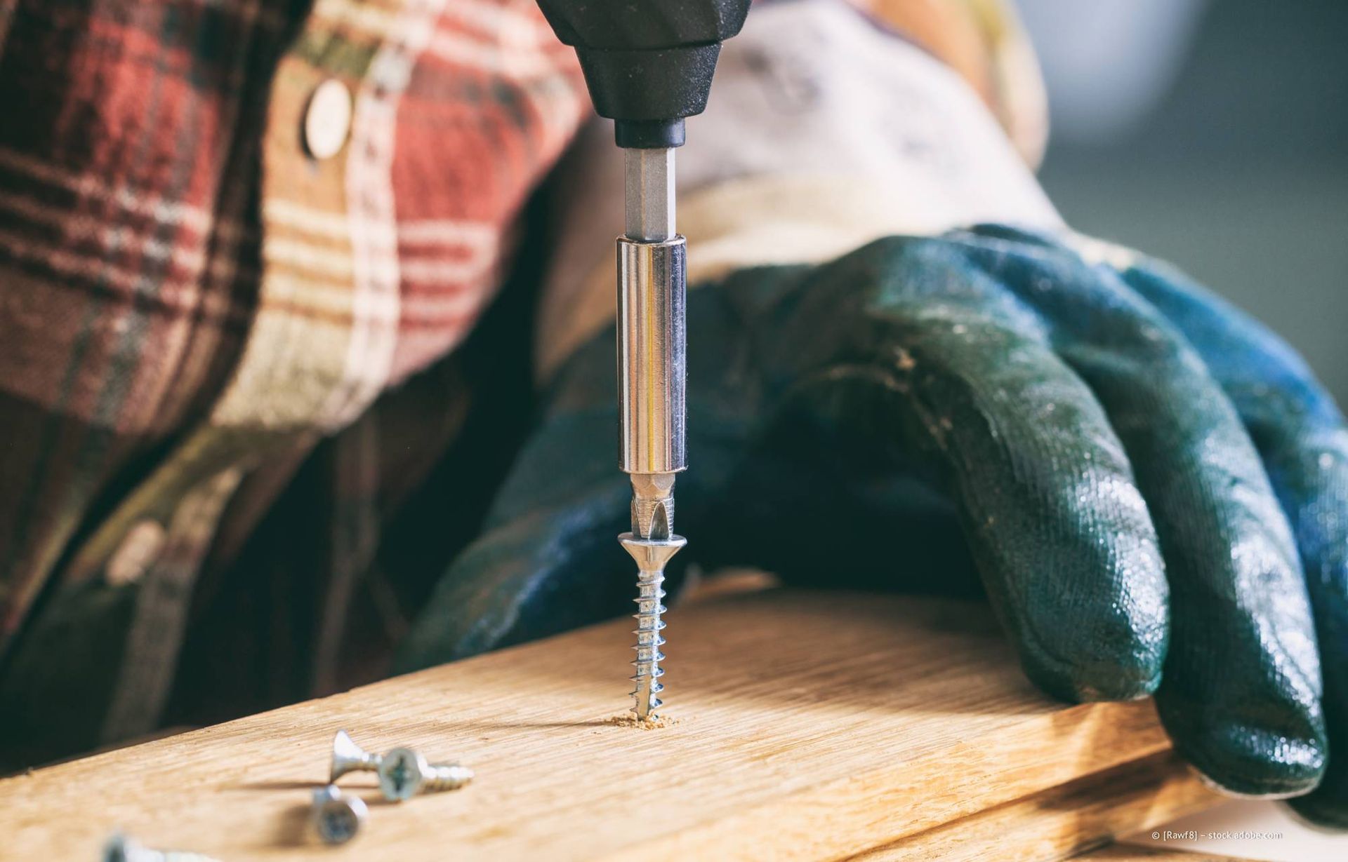 Eine Person mit Arbeitshandschuhen benutzt einen Akkuschrauber, um eine Schraube in ein Stück Holz zu schrauben.