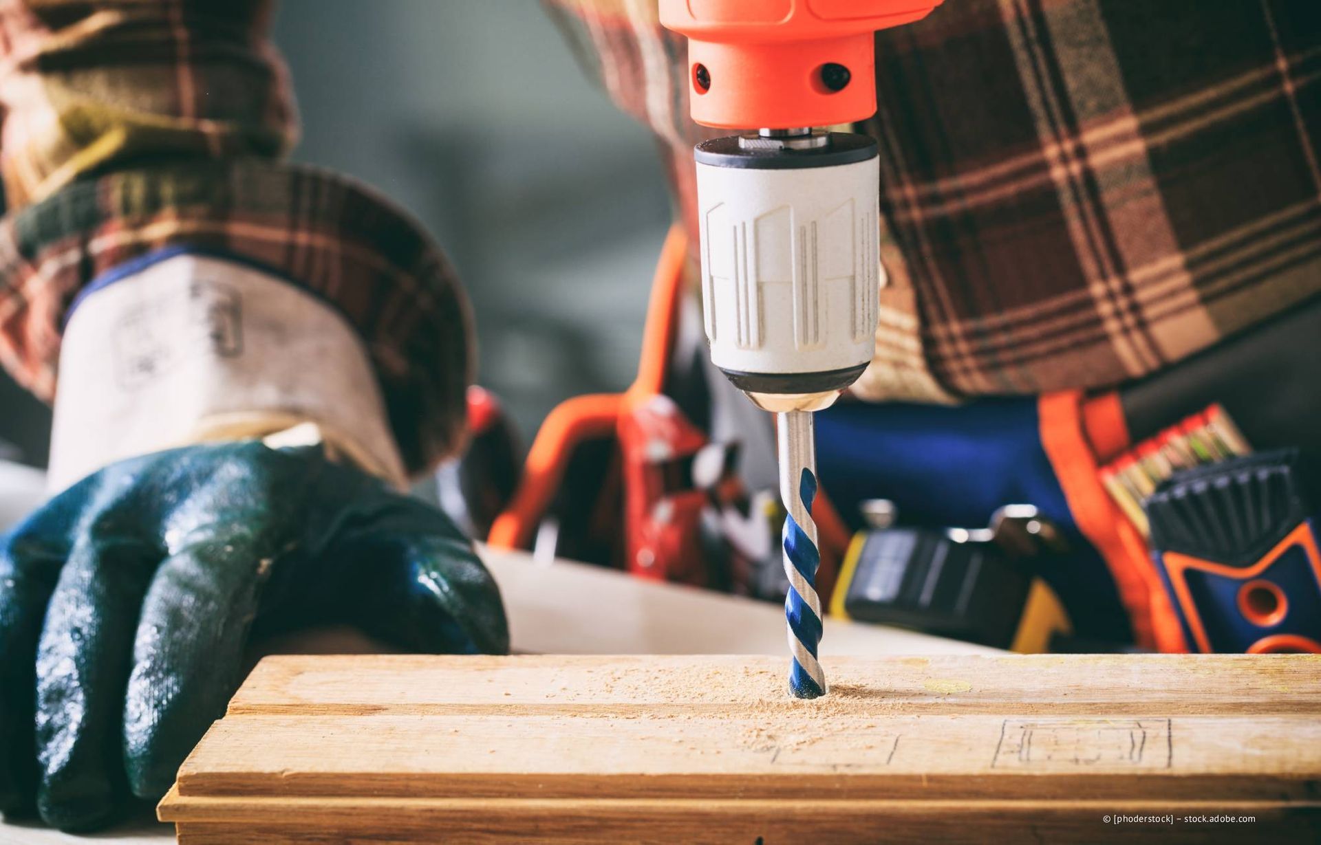 Person, die mit einer Bohrmaschine ein Loch in ein Holzbrett bohrt; orange-weiße Bohrmaschine, mit Schutzhandschuh.