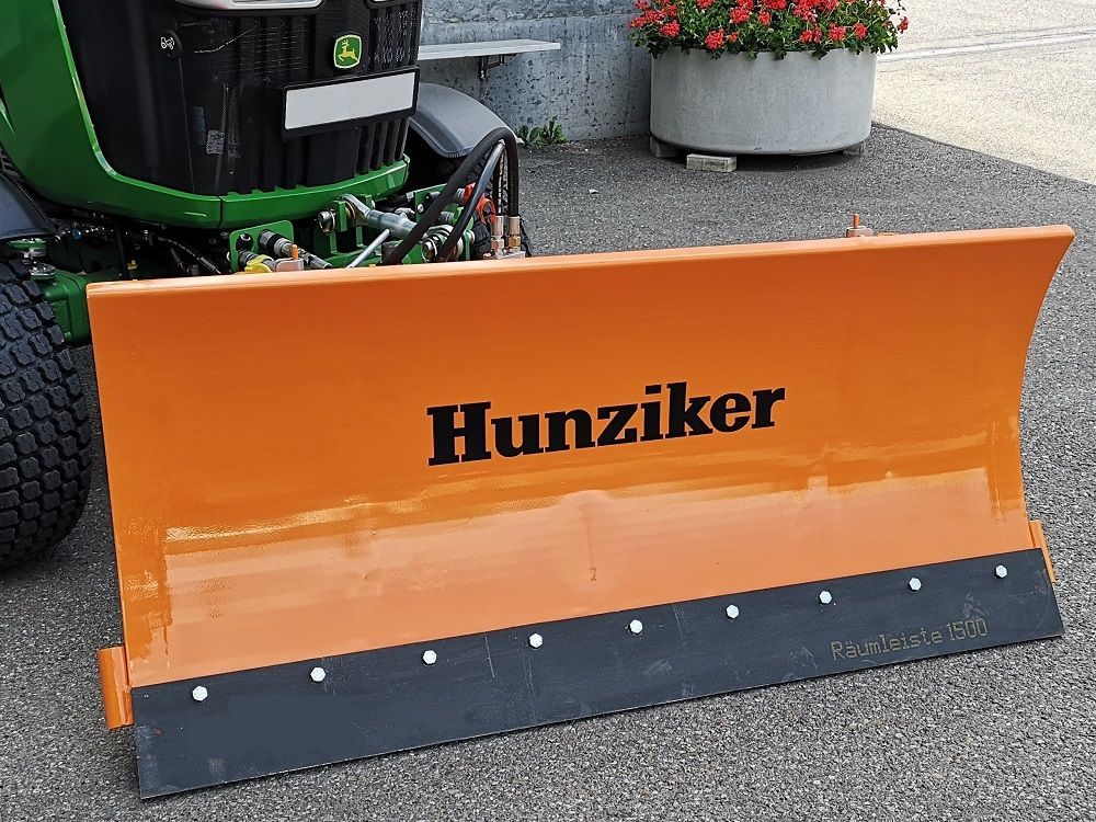 Orangefarbener Hunziker-Schneepflug, befestigt an einem grünen John Deere-Traktor. Der Pflug hat eine schwarze 
