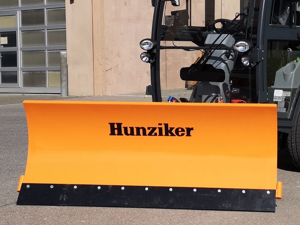 Ein orangefarbener Schneepflug mit dem Namen „Hunziker“ an der Vorderseite eines Fahrzeugs.