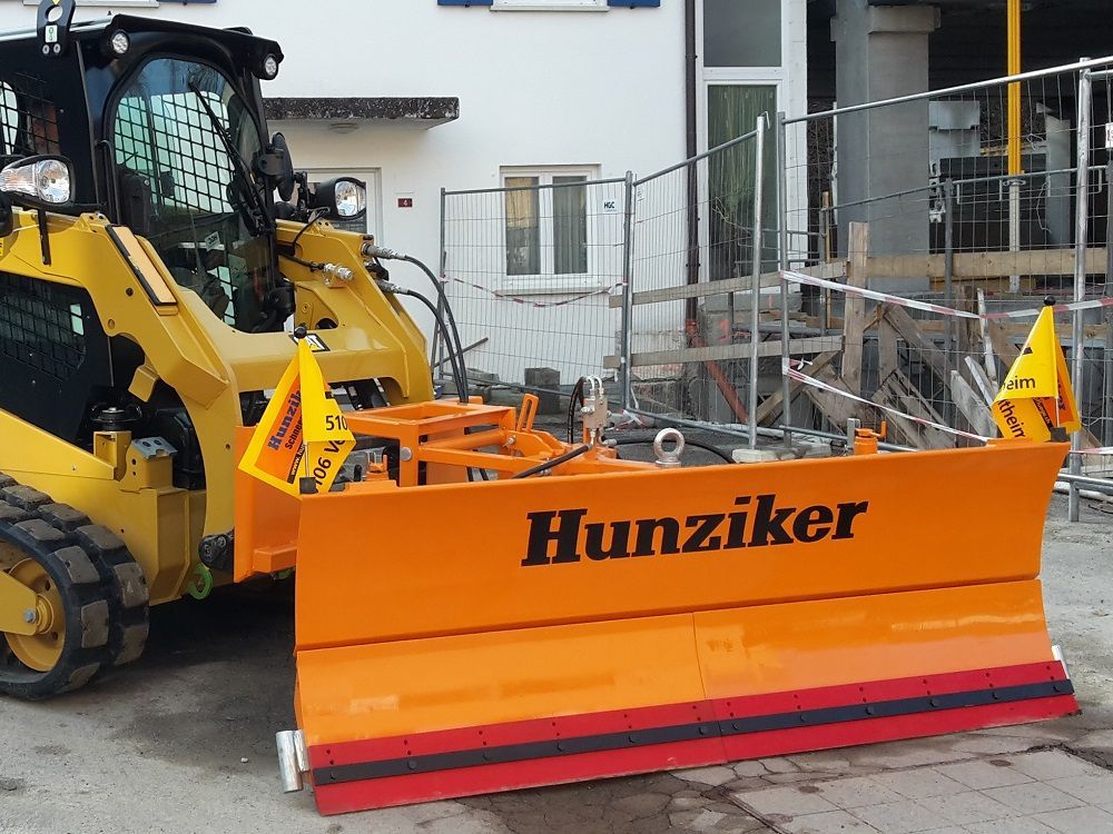 Kompaktlader mit orangefarbenem Schneepflug „Hunziker“ bereitet sich auf die Schneeräumung in einem Baustellenbereich vor.