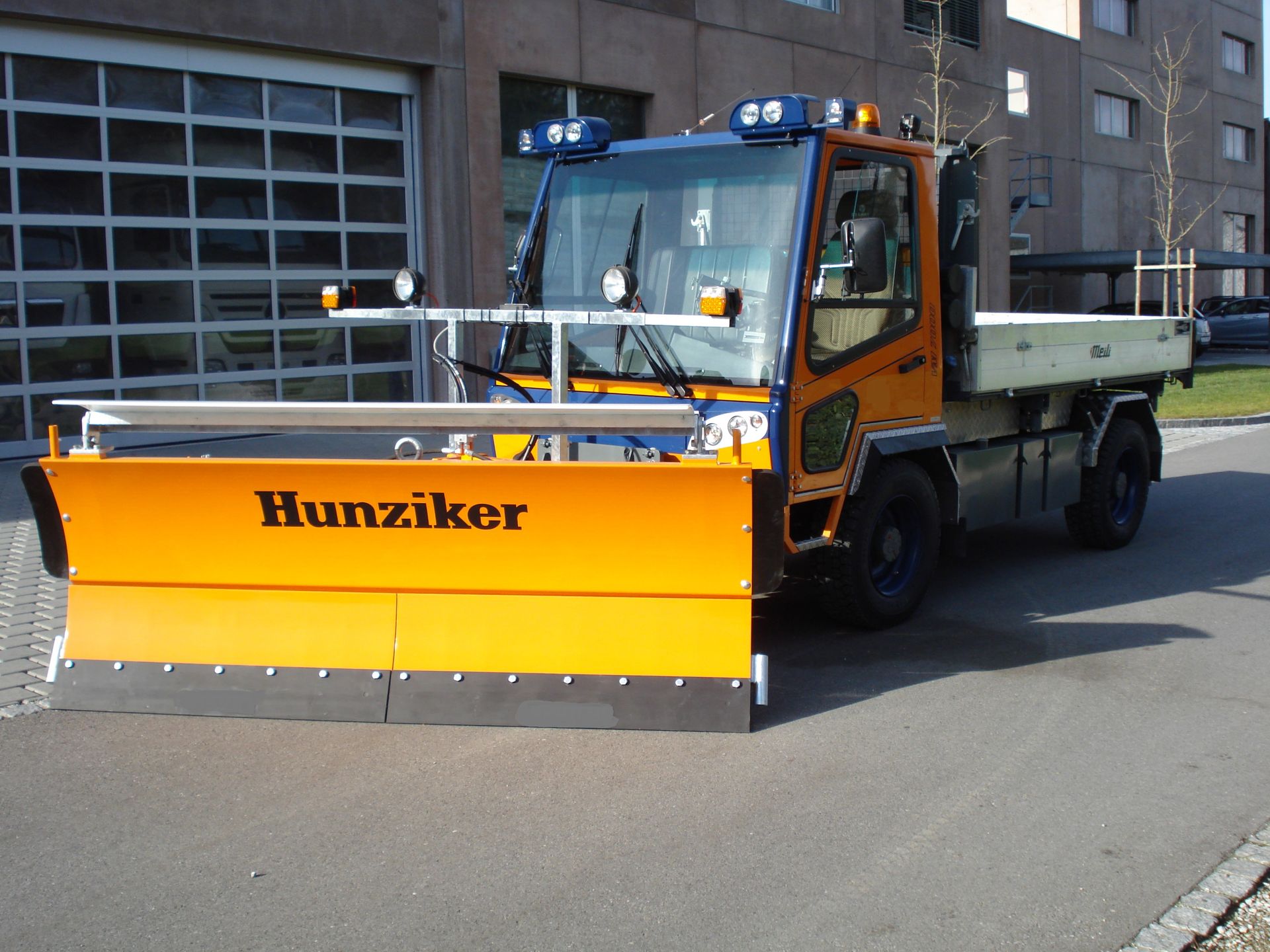 Orange-blauer Hunikzer-Schneepflug auf einer asphaltierten Straße vor einem Gebäude mit Garagentor.