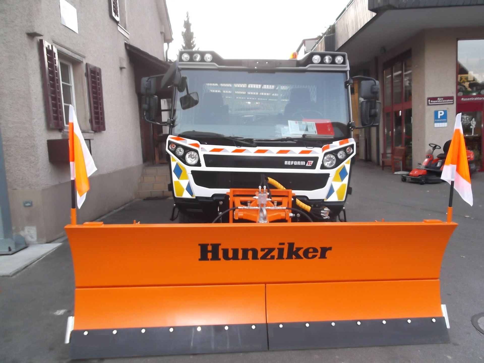 Schneepflug mit dem Namen „Hunziker“ auf der orangefarbenen Klinge, vor einem Gebäude geparkt. Weiß-orangefarbene Flaggen 