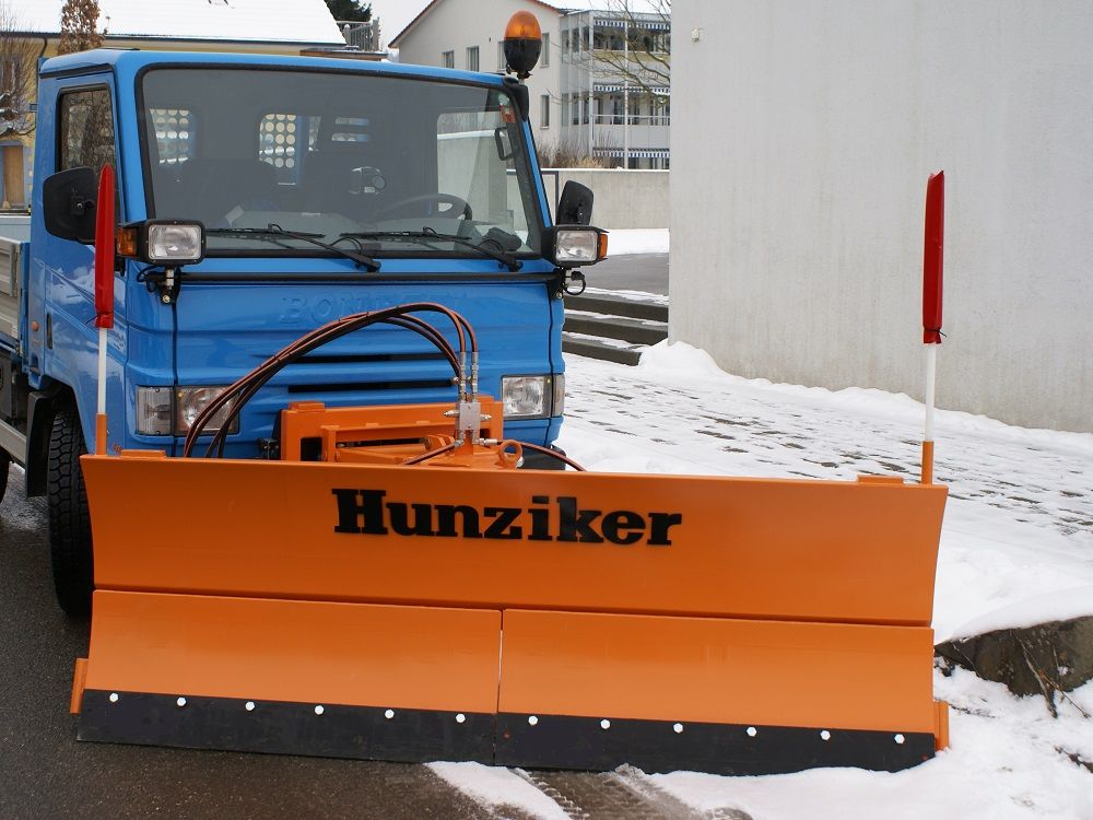 Blauer Schneepflug mit orangefarbener Räumklinge und der Aufschrift „Hunziker“ räumt Schnee von einer Straße.