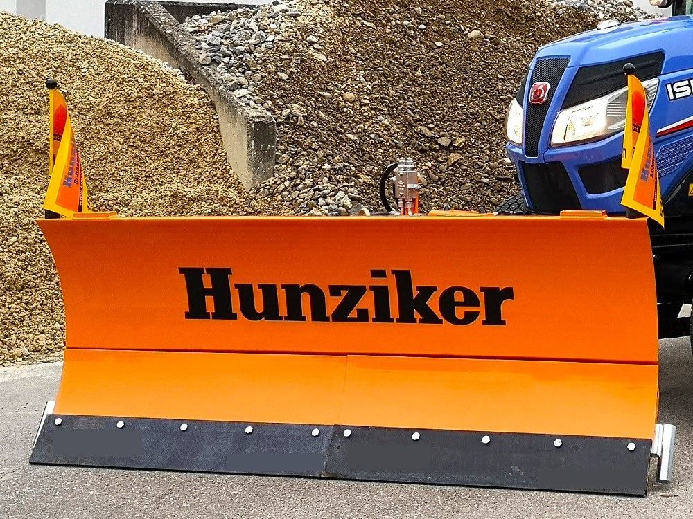Orangefarbener Schneepflug mit der Aufschrift „Hunziker“ an einem blauen Traktor befestigt, im Hintergrund ein Kieshaufen.