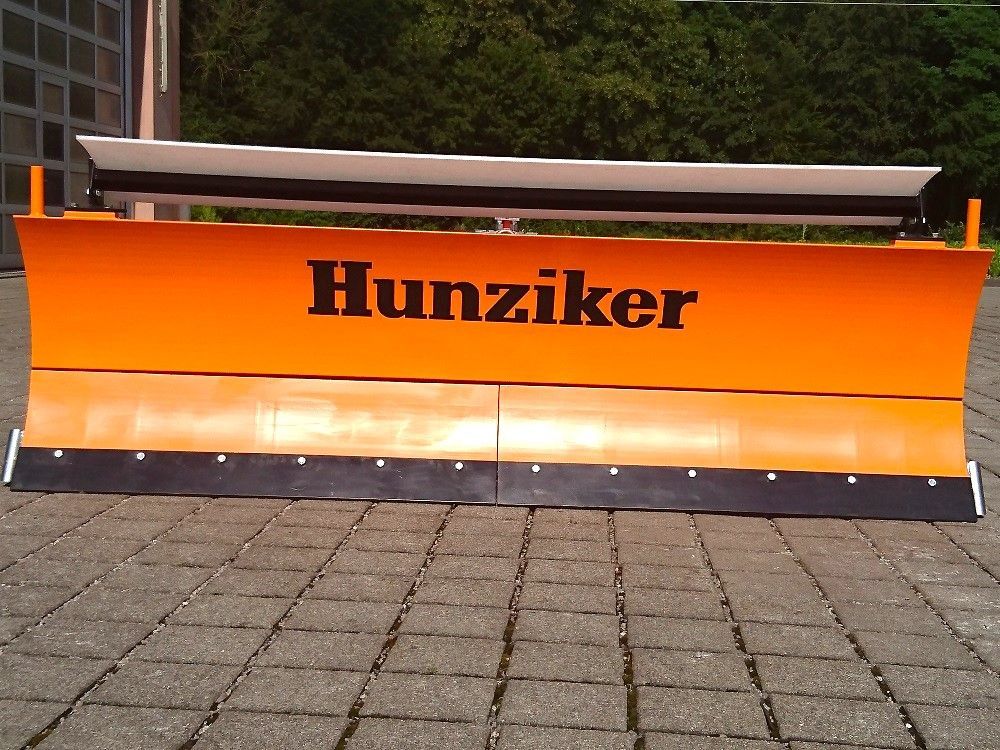 Orangefarbener Hunziker-Schneepflug mit schwarzer Gummiklinge auf einer gepflasterten Fläche.