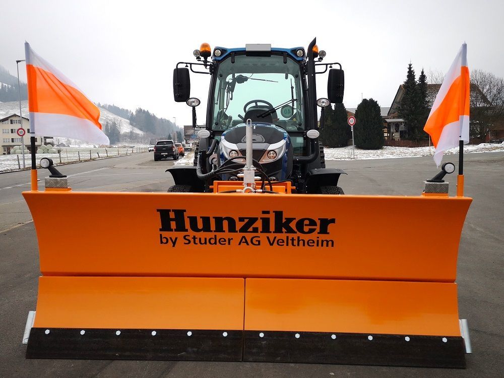 Orangefarbener Schneepflug an einem blauen Traktor mit orange-weißen Fahnen. Der Text auf dem Pflug lautet 