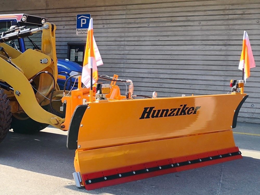Orangefarbener Hunziker-Schneepflug an einem gelben Lader mit orange-weißen Fahnen. Der Pflug ist vor einem Gebäude geparkt.