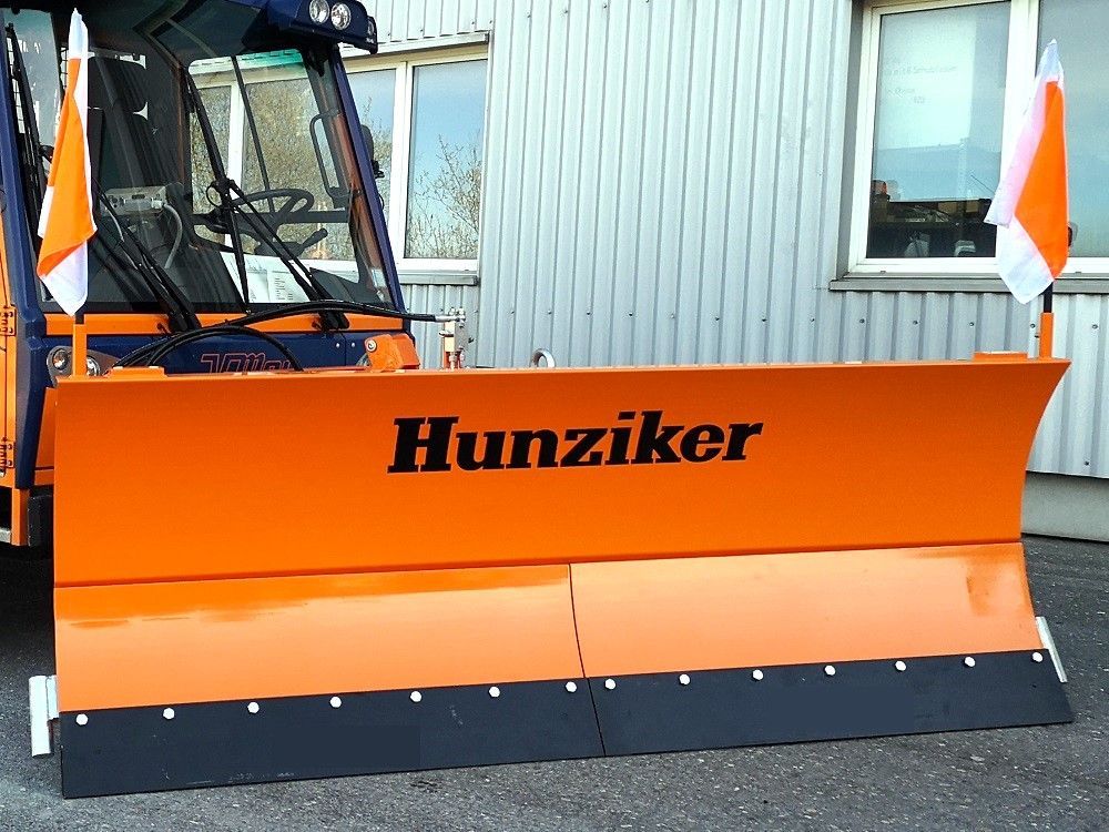 Orangefarbener Schneepflug mit „Hunziker“-Branding, im Freien mit weiß-orangen Fahnen geparkt.