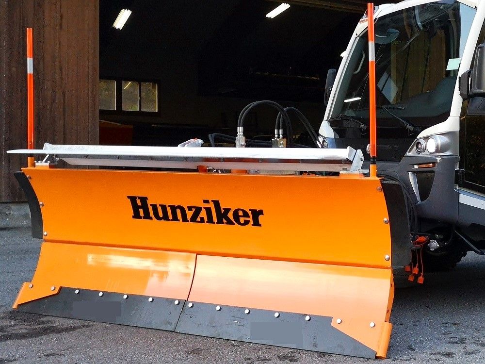 Orangefarbener Schneepflug an einem weißen LKW, auf dem das „Hunziker“-Logo zu sehen ist.
