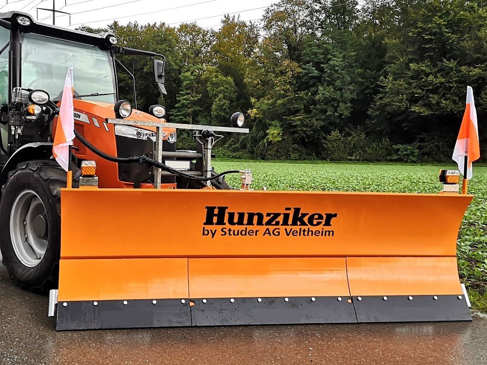 Orangefarbener Hunziker-Schneepflug, befestigt an einem Traktor, mit Fahnen, auf einer asphaltierten Straße.