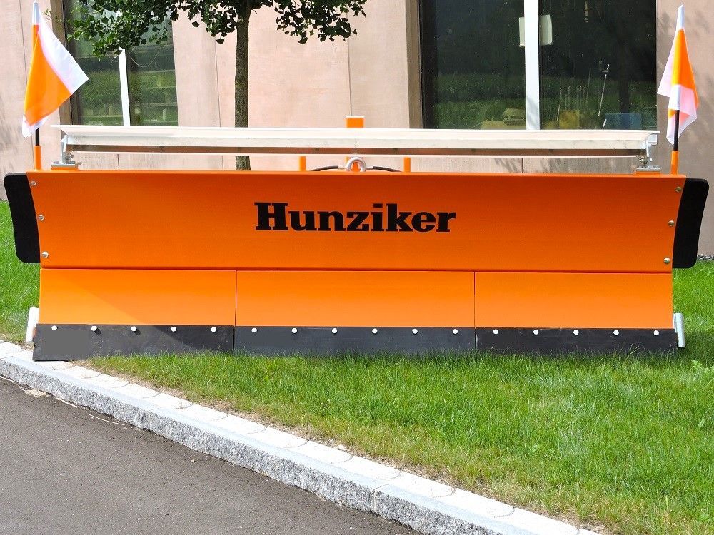 Orangefarbener Schneepflug mit dem Namen „Hunziker“, geparkt am Rand einer Rasenfläche mit zwei orange-weißen Fahnen.