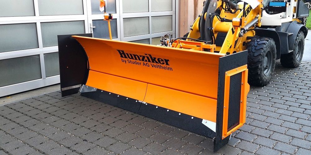 Ein orange-schwarzer Schneepflug ist an einem gelb-weißen Traktor befestigt. Der Schneepflug steht auf einer gemauerten Fläch