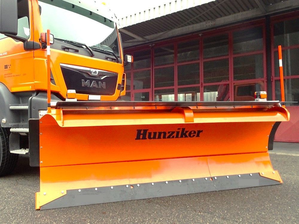 Orangefarbener Schneepflug, befestigt an einem dazu passenden orangefarbenen MAN-LKW, der vor einem Gebäude