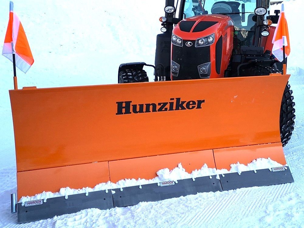 Orangefarbener Schneepflug an einem Traktor beim Schneeräumen. Auf dem Pflug steht der Name „Hunziker“.