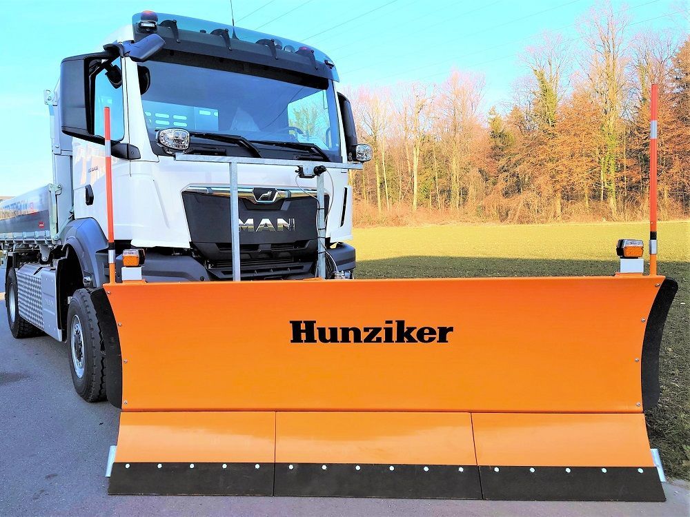 Orangefarbener Schneepflug an einem MAN-LKW, bereit zum Räumen einer Straße. Der Markenname „Hunziker“ ist sichtbar.