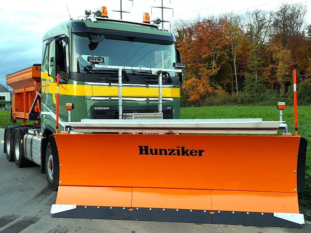 Orangefarbener Schneepflug mit der Aufschrift „Hunziker“ auf einer Straße. Der LKW ist mit Herbstlaub im Hintergrund geparkt.