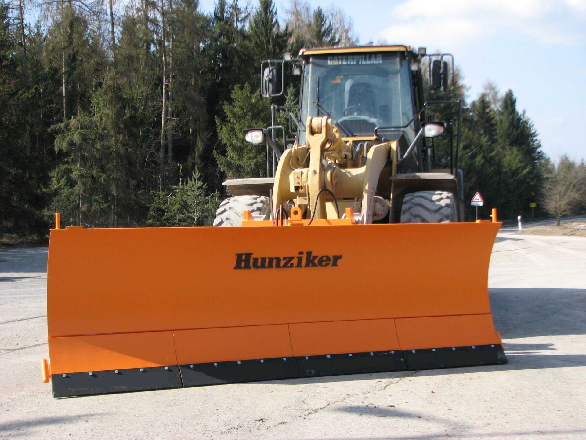 Gelber Frontlader mit orangefarbener Schneeräumschaufel auf einer befestigten Fläche in der Nähe von Bäumen. Auf der 