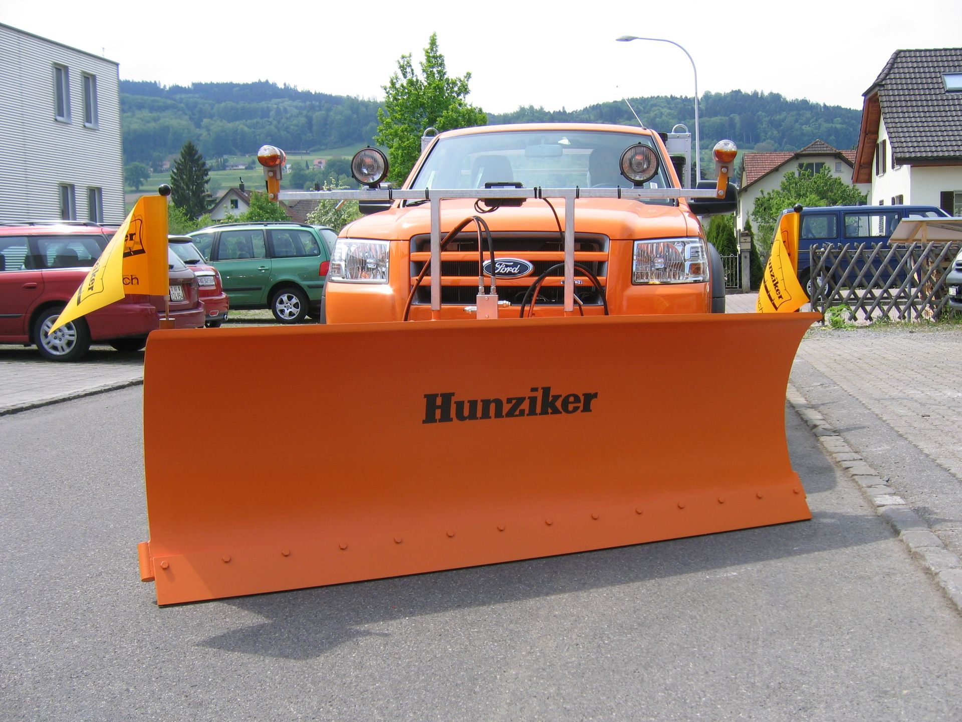 Orangefarbener Schneepflug, befestigt an einem orangefarbenen Ford-Truck, geparkt auf einer asphaltierten Straße. Auf der Kli