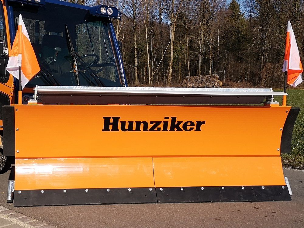 Ein orangefarbener Hunziker-Schneepflug mit einer schwarzen Klinge, montiert auf einem Fahrzeug, mit orangefarbenen und 