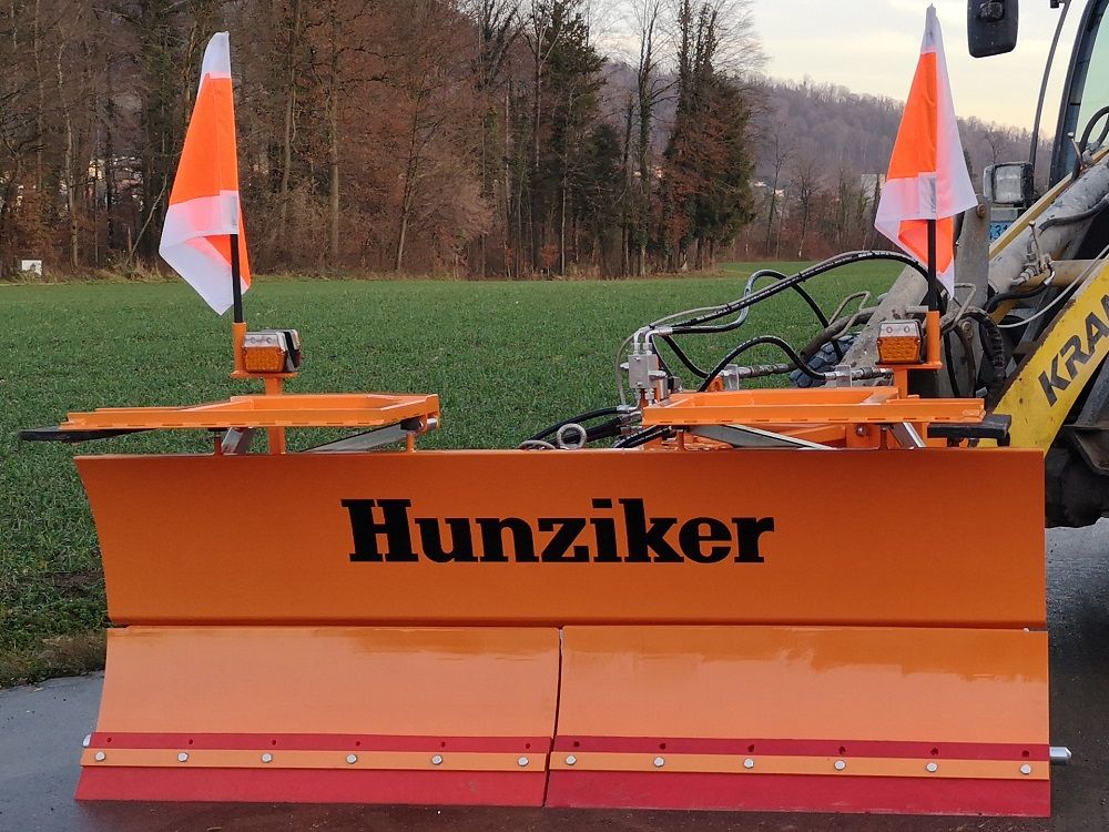Orangefarbener Hunziker-Schneepflug mit angebrachten Fahnen, auf Asphalt vor einer grünen Wiese und Bäumen.