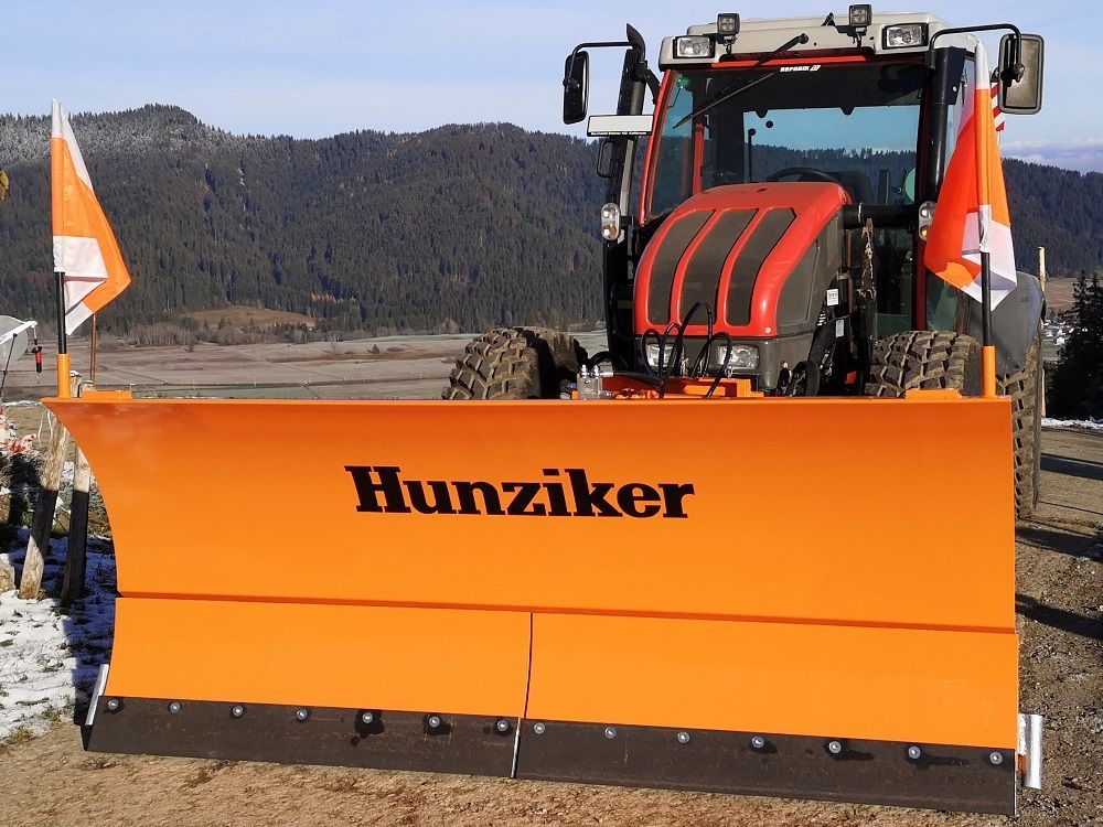 Orangefarbener Schneepflug an einem roten Traktor, auf dem der Name „Hunziker“ zu sehen ist. Am Fahrzeug sind orangefarbene F
