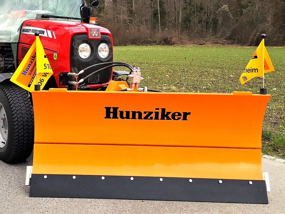 Auf einer asphaltierten Straße parkt ein roter Traktor mit einem orangefarbenen Schneepflug, der den Namen „Hunziker“ trägt, 