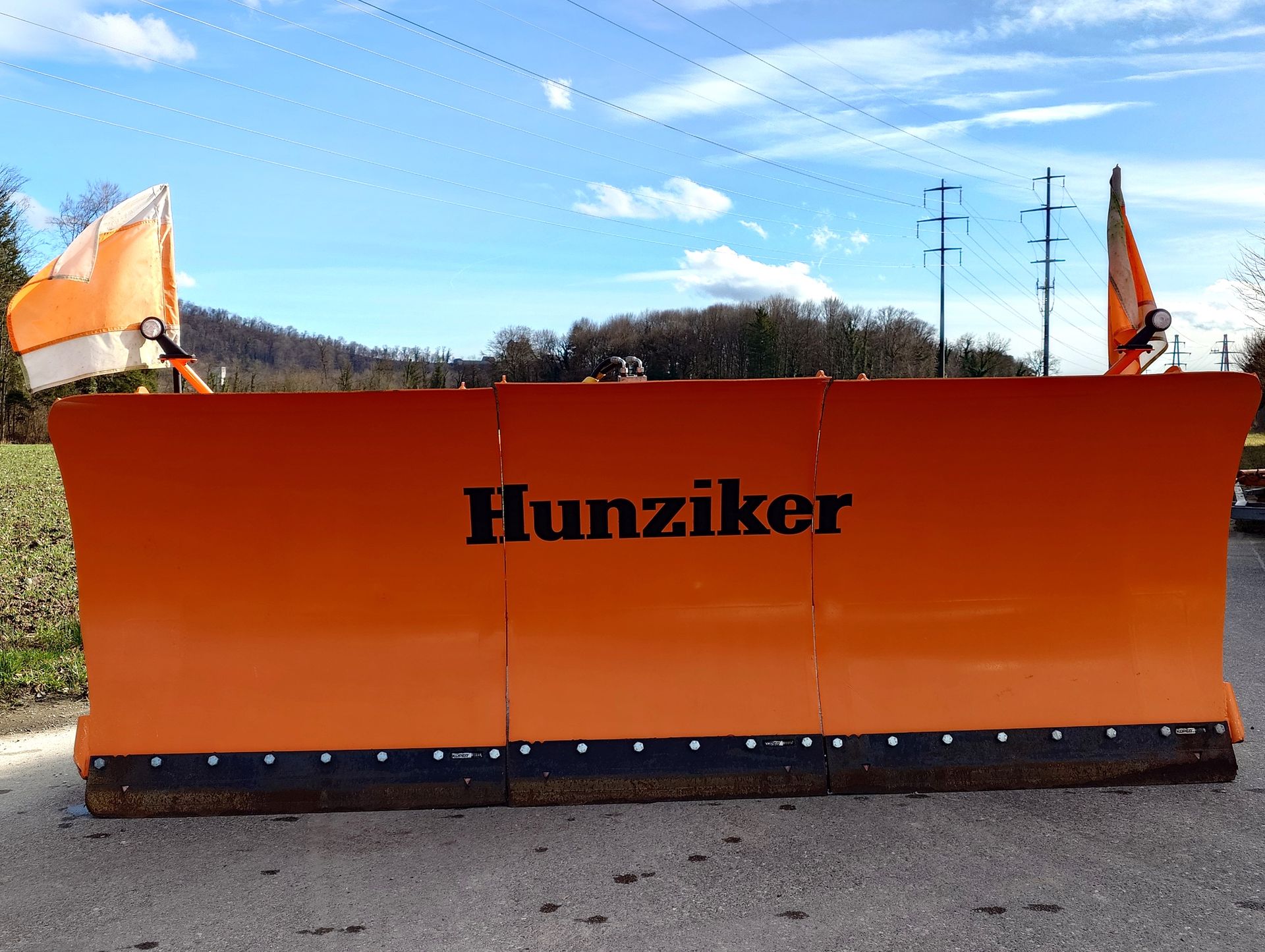 Orangefarbener Hunziker-Schneepflug auf einer Straße mit Sicherheitsfahnen.