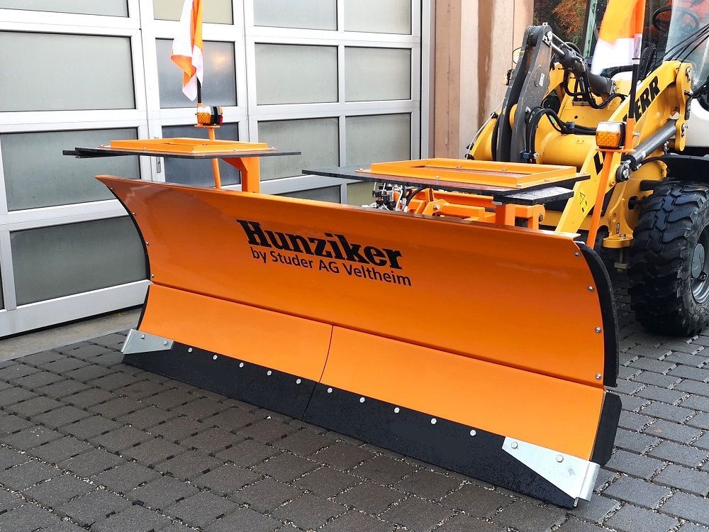 Orangefarbener Schneepflug an einem gelben Lader, geparkt vor einem Gebäude mit weißer Tür. Auf dem Pflug 