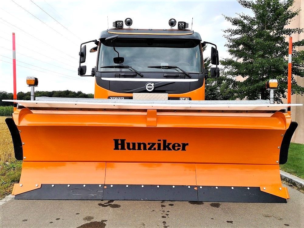 Orangefarbener Hunziker-Schneepflug, an einem Volvo-LKW befestigt, bereit für die Straßenräumung im Winter.
