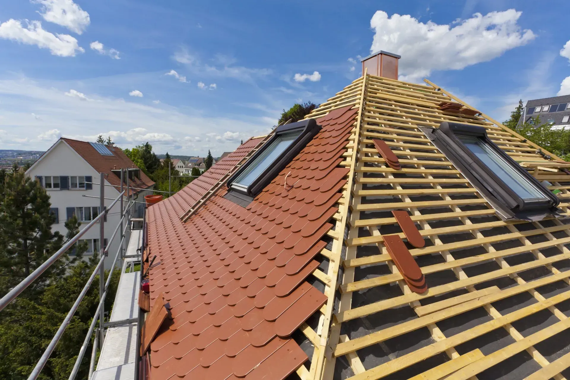 Toiture en construction avec tuiles rouges, liteaux de bois apparents et puits de lumière sous un ciel bleu