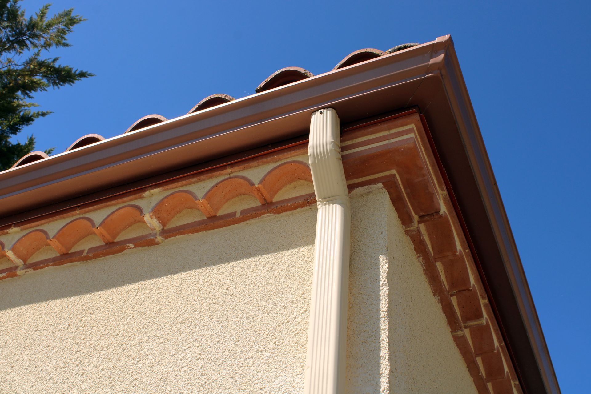 Vue en contre-plongée d'un coin de maison en stuc beige avec des tuiles rouges et une gouttière blanche sur fond de ciel bleu