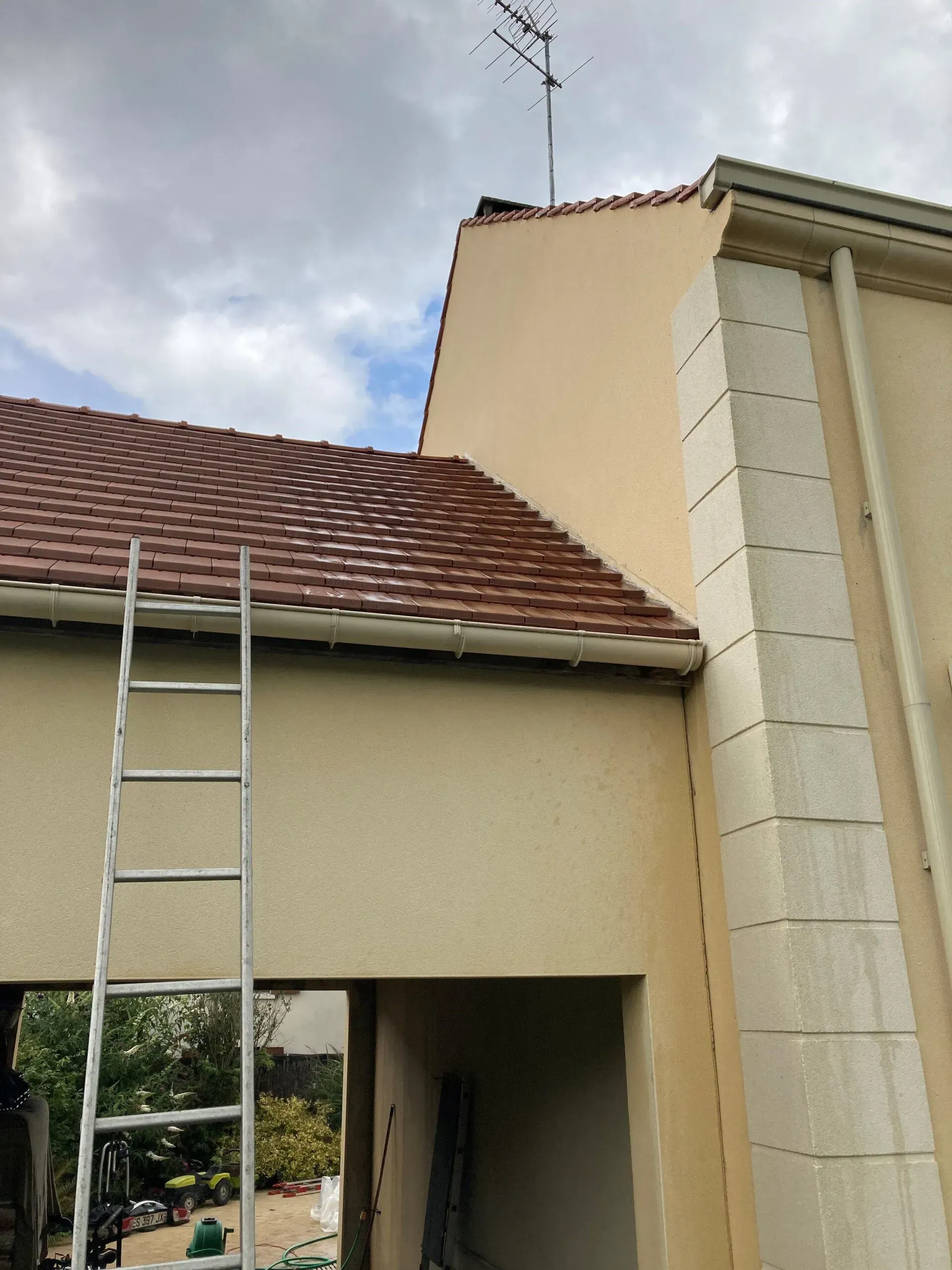 Extérieur de la maison avec toit, échelle contre l'avant-toit et entrée partiellement visible sous un ciel nuageux
