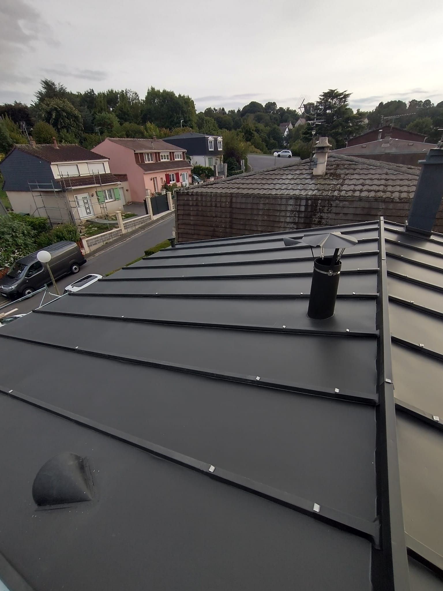 Toit avec panneaux métalliques sombres, aérations et maisons voisines sous un ciel nuageux
