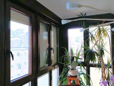 Balcón soleado con ventanas de marco marrón y macetas, con vistas al paisaje urbano. Se ve una lámpara moderna.