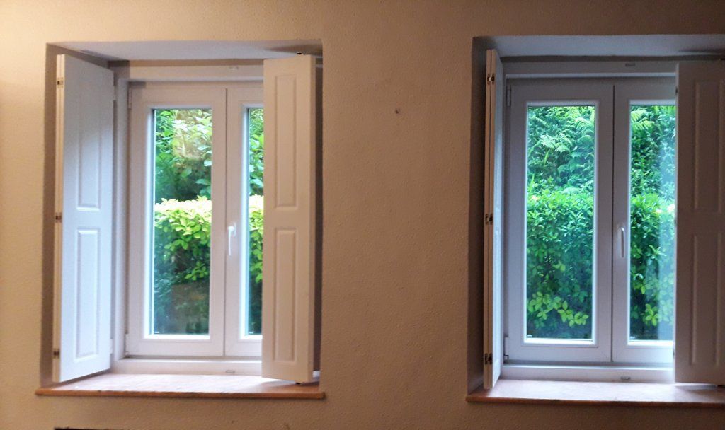 Dos ventanas con marco blanco y contraventanas blancas abiertas. A través de los cristales, enmarcadas por una pared color crema, se ve una vegetación verde.