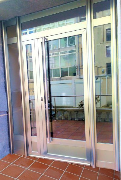 Puerta de cristal con marco plateado y ventanas laterales que reflejan un edificio. Suelo exterior de baldosas marrones.