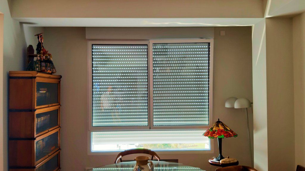 Una ventana con mosquitera y persianas está flanqueada por una estantería y una mesa con una lámpara. Las paredes son beige.