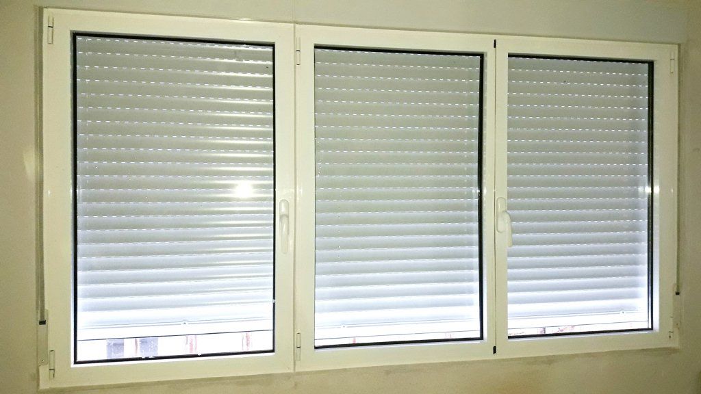 Tres ventanas con marco blanco y persianas cerradas sobre una pared de color claro.