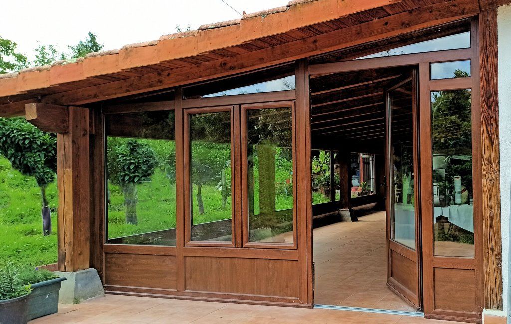 Entrada de cristal con marco de madera marrón y puerta abierta. Tiene techo de tejas y da a un sendero con vegetación.