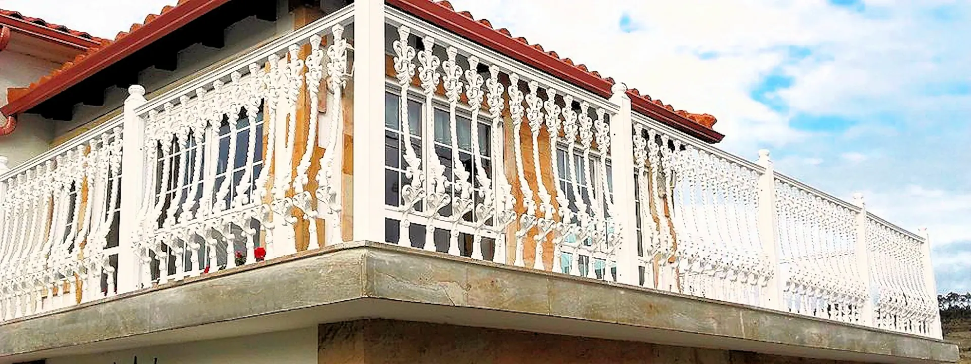 Balcón de una casa con barandilla decorativa blanca y techo de tejas rojas contra un cielo nublado.