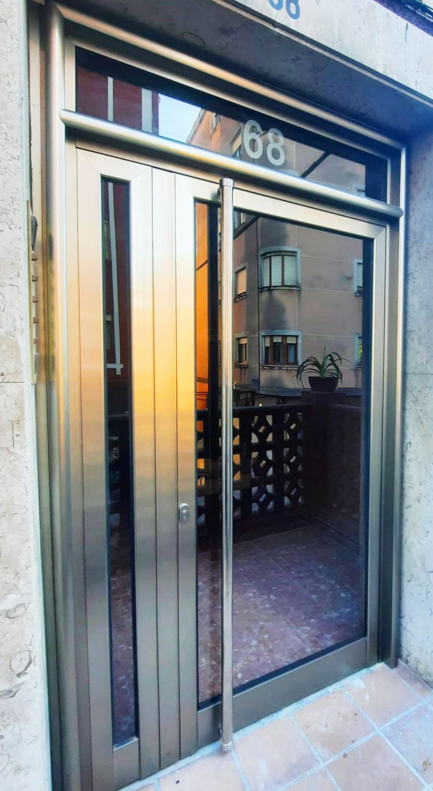Una puerta moderna de vidrio y metal con el número 68 encima. Los reflejos muestran un balcón y un vistazo a un edificio de apartamentos.