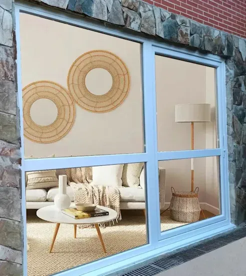 Puertas corredizas de vidrio que revelan una sala de estar con muebles de colores claros, una mesa de café y una decoración de pared tejida, frente a un exterior de piedra.