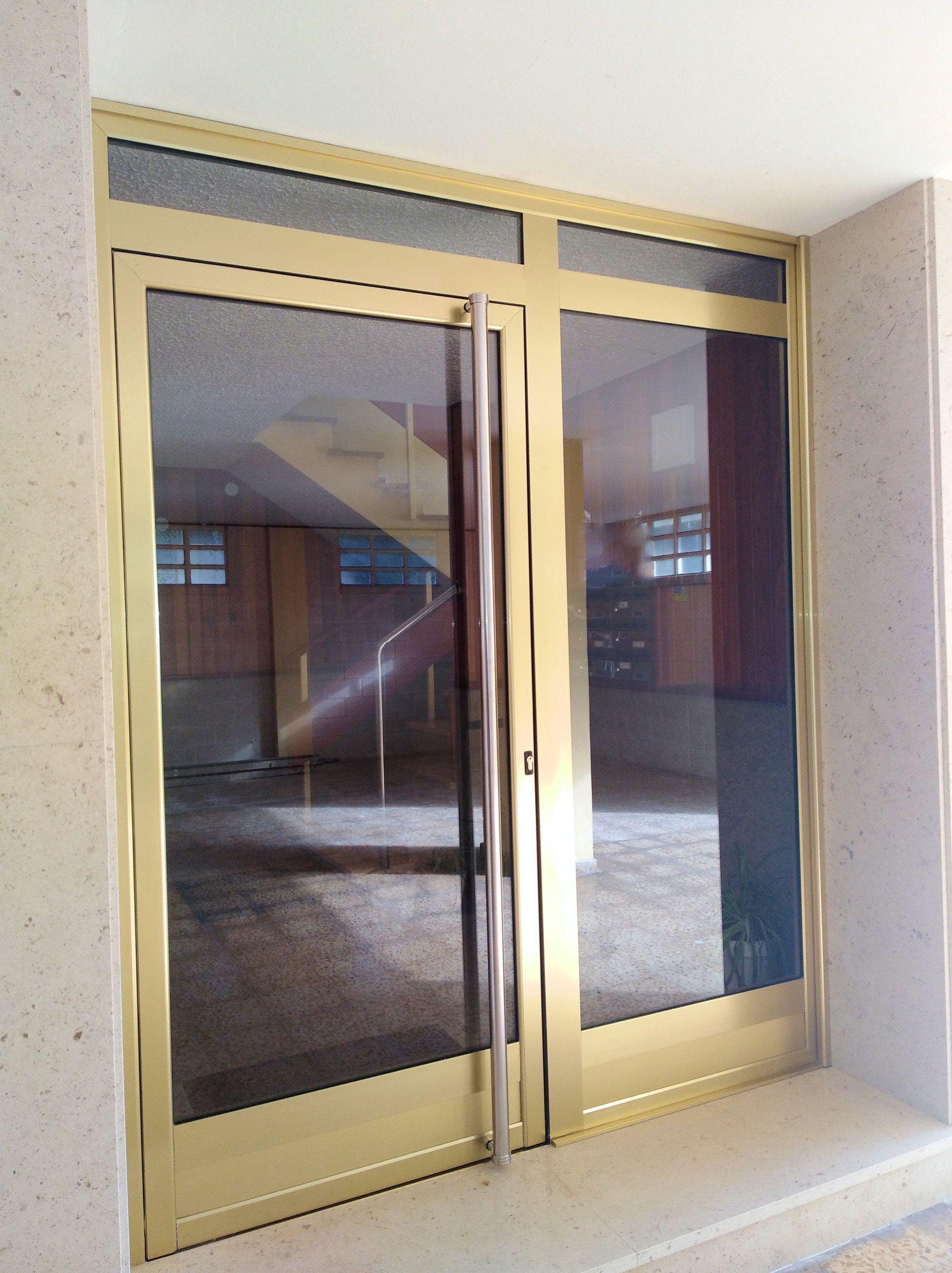 Puertas dobles de cristal con marcos dorados que reflejan un pasillo con escalera. Paredes beige claro rodean la entrada.
