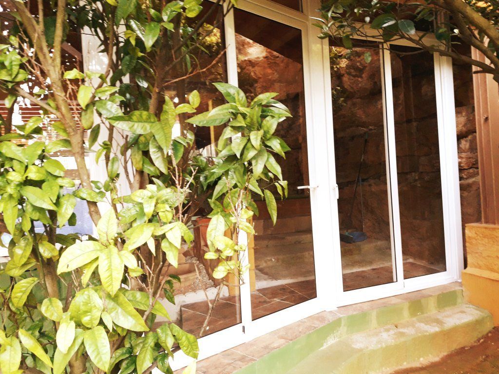 Puertas de cristal con marcos blancos, rodeadas de exuberante vegetación. Entrada a un edificio visible, con escaleras en el interior.